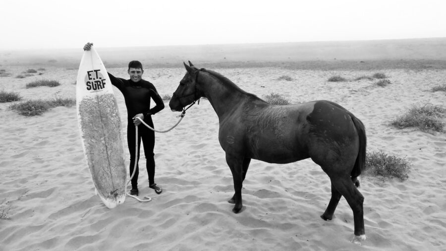 Black & white image of a man holding a surfboard standing with a horse to show a different horsemanship view